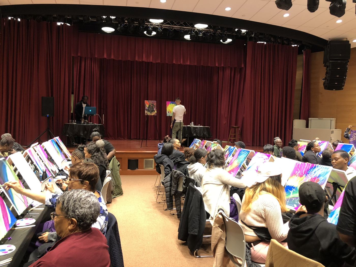 A community auditorium filled with painters at easels while a DJ and live artist work from the stage