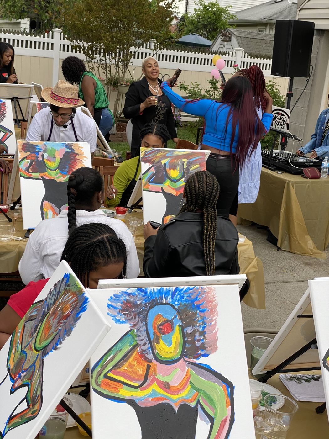An outdoor backyard Music Paint Sip party with a DJ table, rows of easels, and guests painting colorful portrait canvases