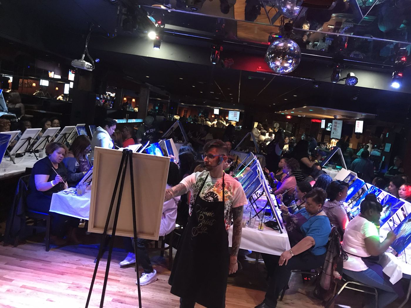 A packed Music Paint Sip night at a club venue, guests painting at long tables under a disco ball while the artist guides from center floor