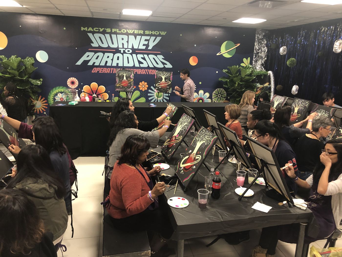 A full room of guests painting fairy and moon canvases at the Macy's Flower Show, guided by the Music Paint Sip artist beneath the Flower Show backdrop